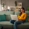 Smiling caucasian woman using laptop while sitting on a mint couch at home. Happy female shopping or chatting online in social network, typing blog, freelancer working on a project.